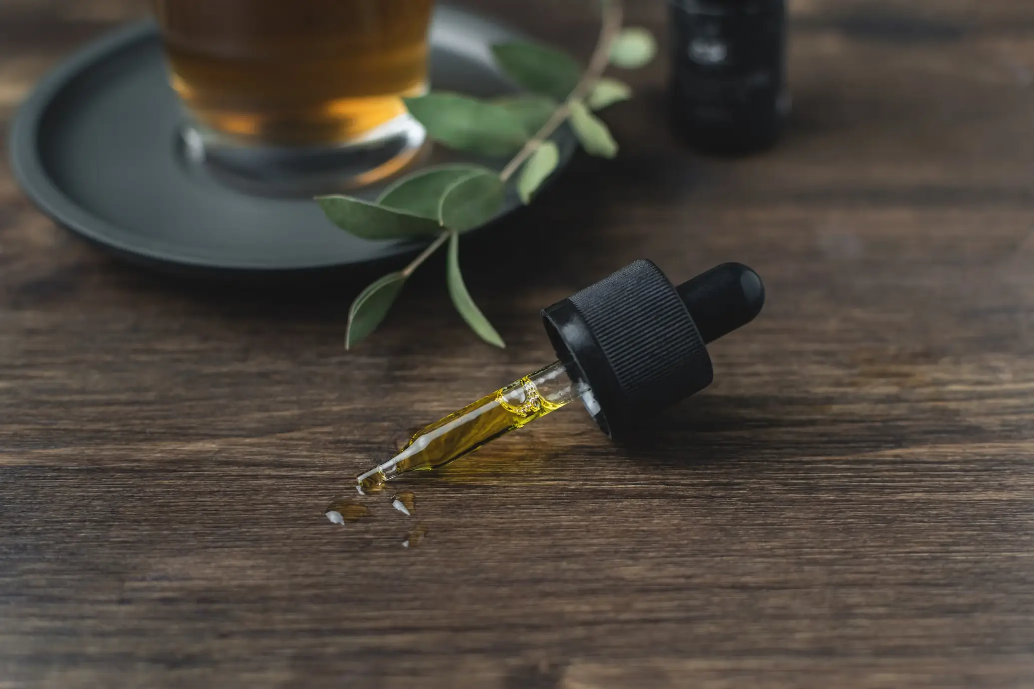 A dropper with essential oil resting on a wooden table, next to a green branch and a cup of tea in the background.